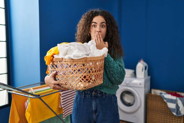 Woman holding a laundry basket, looking surprised in a laundry room, symbolizing common laundry mistakes.